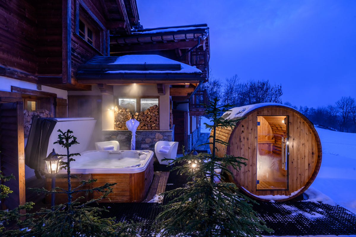 A wooden hot tub and a round wooden sauna are set outside a log cabin in a snowy winter landscape at dusk, with snow-covered ground, evergreen trees, and a blue evening sky.