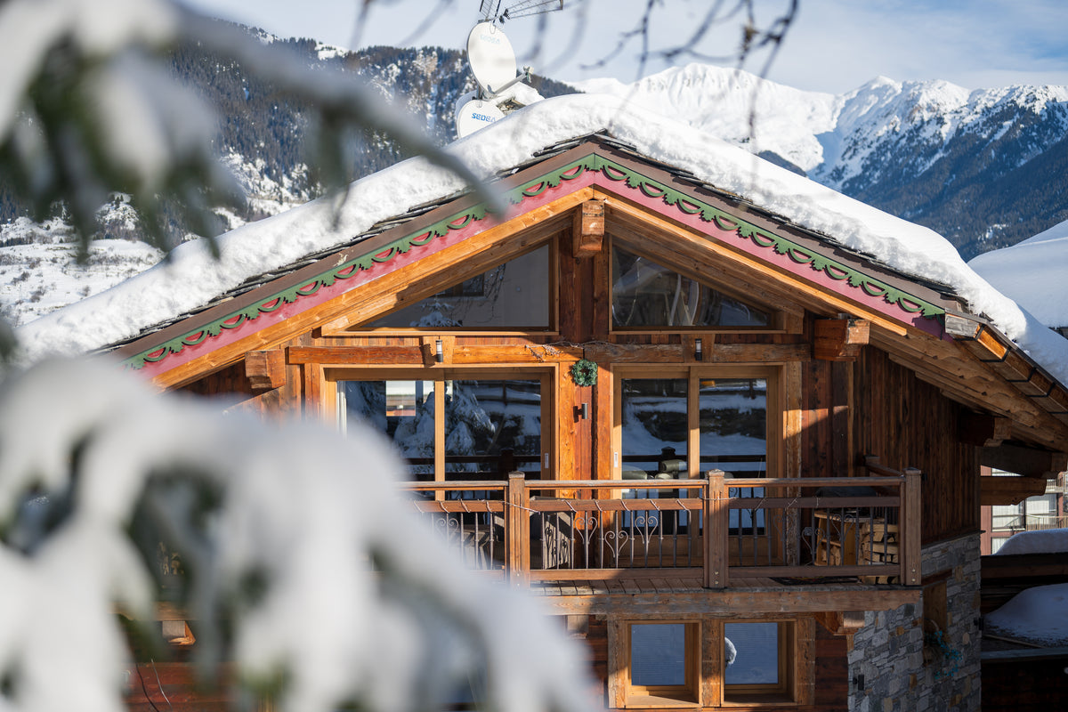 A rustic wooden chalet with snow-covered roof and balcony, set against a snowy mountain landscape with blurred tree branches in the foreground.