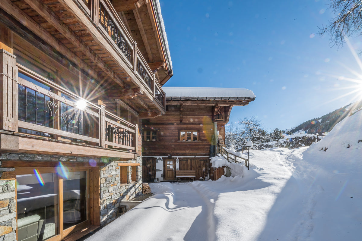 A luxury alpine chalet with wooden balconies and stone walls, surrounded by deep snow under a bright blue sky with sun rays.