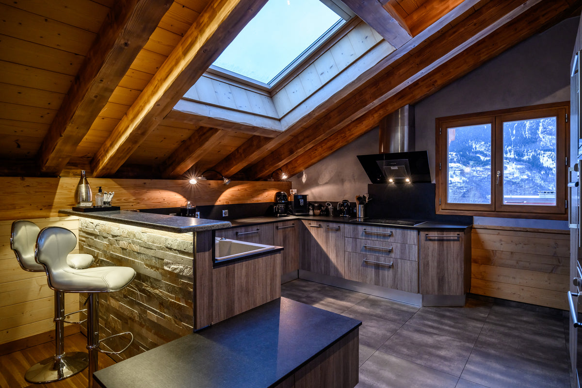 A modern kitchen installed in an attic space with exposed wooden beams and a skylight. The kitchen features light wood cabinetry, dark countertops, a stone-tile backsplash, and a matching island.