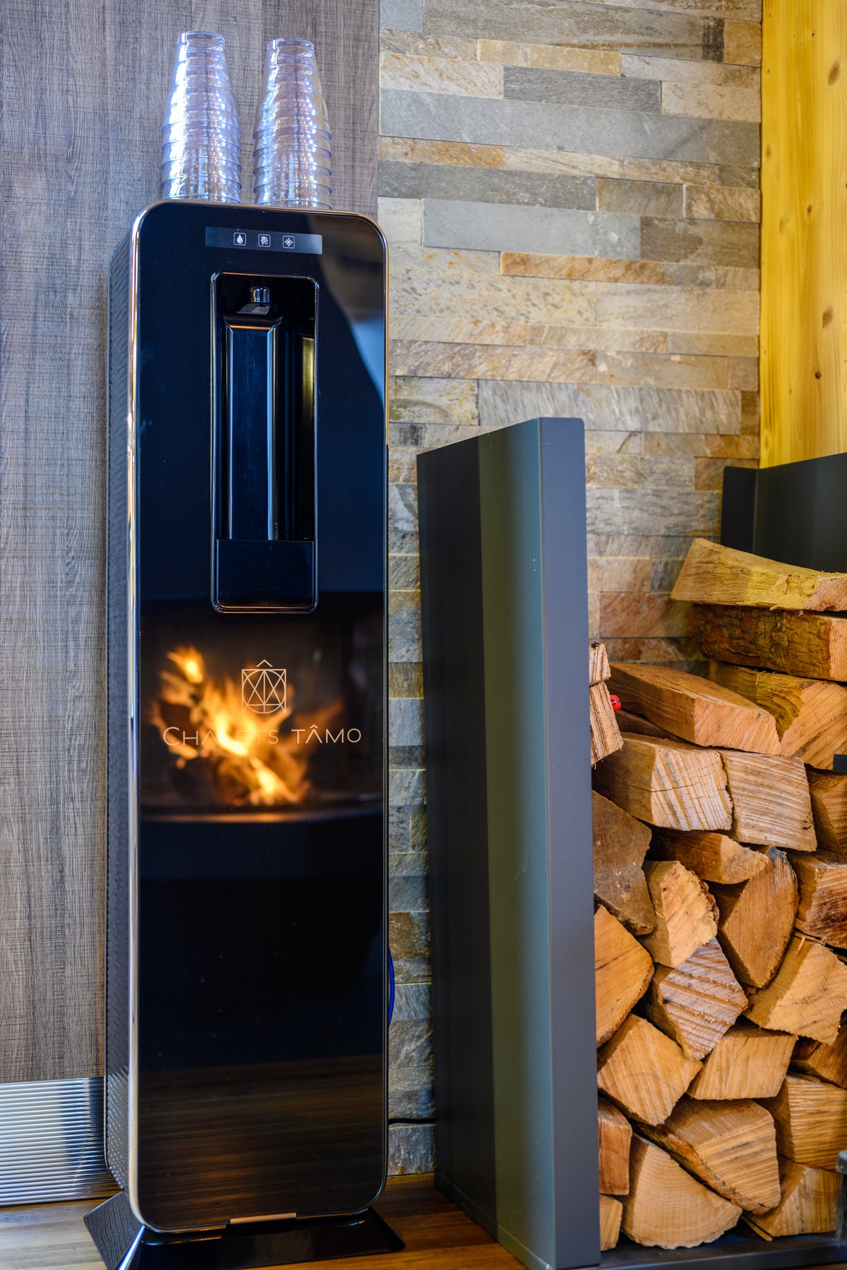 A modern black glass water dispenser with a built-in fire element, shown next to a stack of firewood and a tall gray side table in a rustic interior setting.