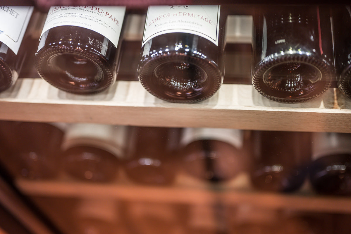 Close-up of several dark glass wine bottles with white labels resting on a wooden shelf in a wine cellar or refrigerator.