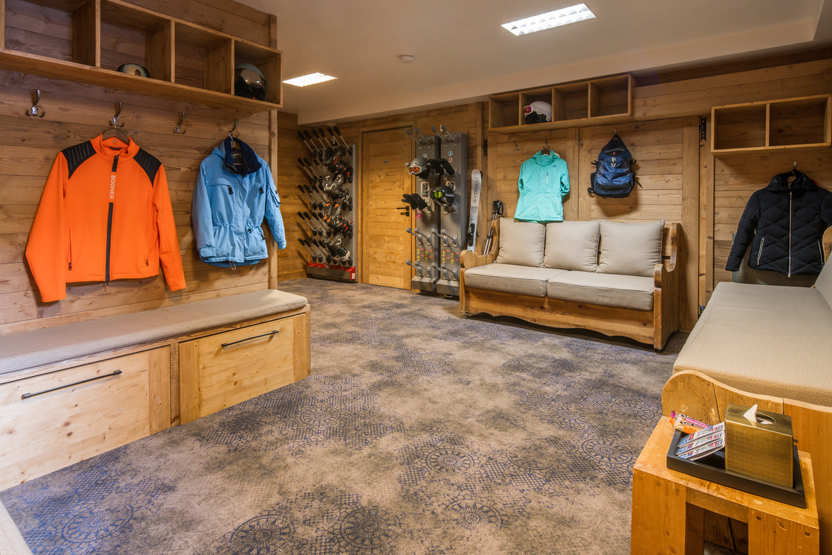 A ski lodge or alpine changing room with wood-paneled walls, lockers, a bench, and several jackets hanging on display. Visible jackets include an orange zip-up, a light blue jacket, a turquoise top, and a dark navy puffer jacket, along with backpacks and ski gear.