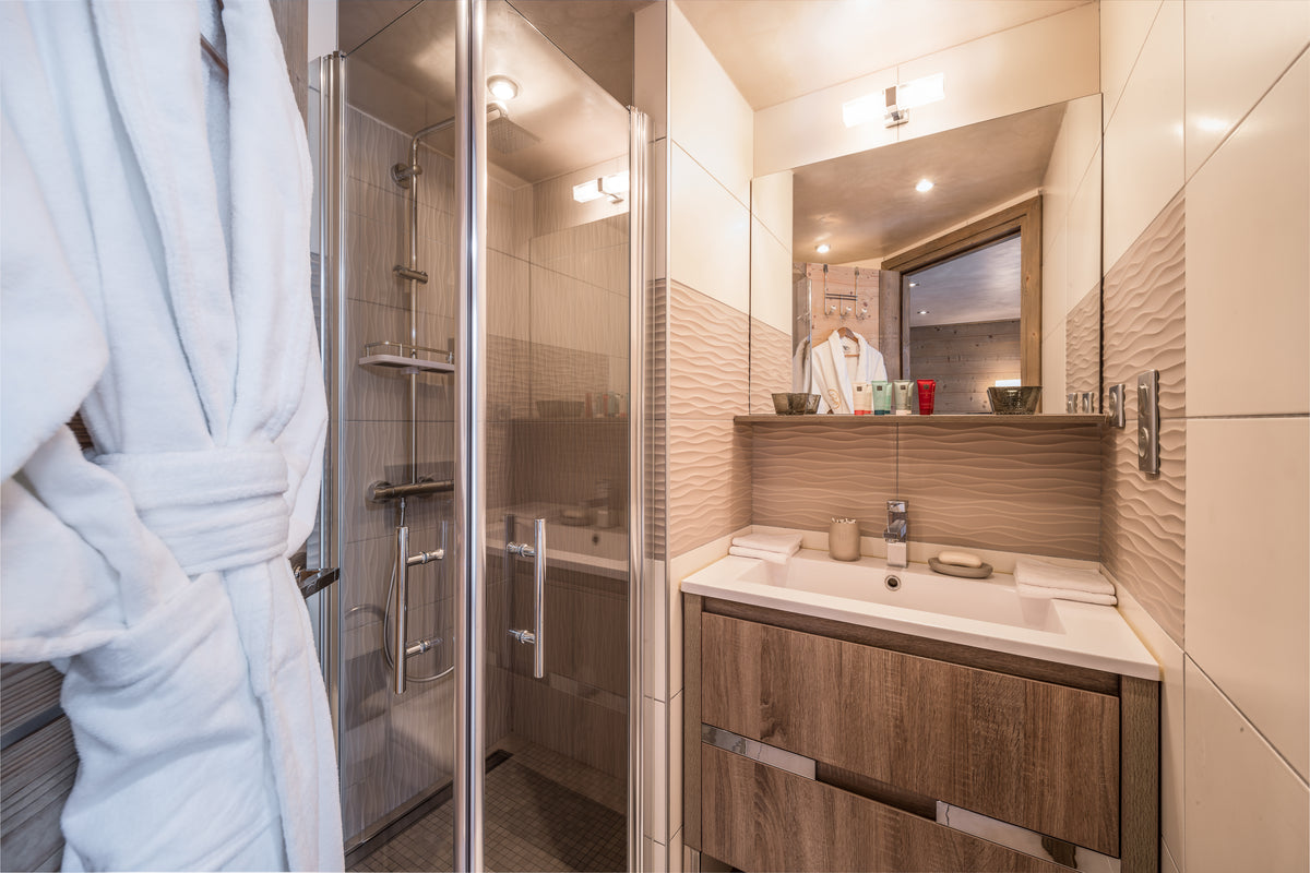 A modern bathroom interior with a glass shower enclosure on the left, a wood-look vanity with a white sink countertop, and a large mirror above. A light blue bathrobe is draped in the foreground on the left side.