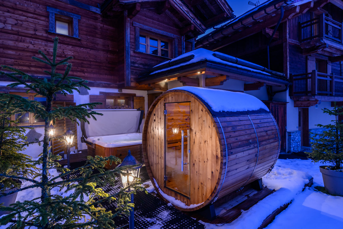A wooden outdoor sauna with a snow-covered barrel-shaped exterior and glowing warm lights, set in front of a rustic alpine chalet with snow on the ground and trees in the foreground.