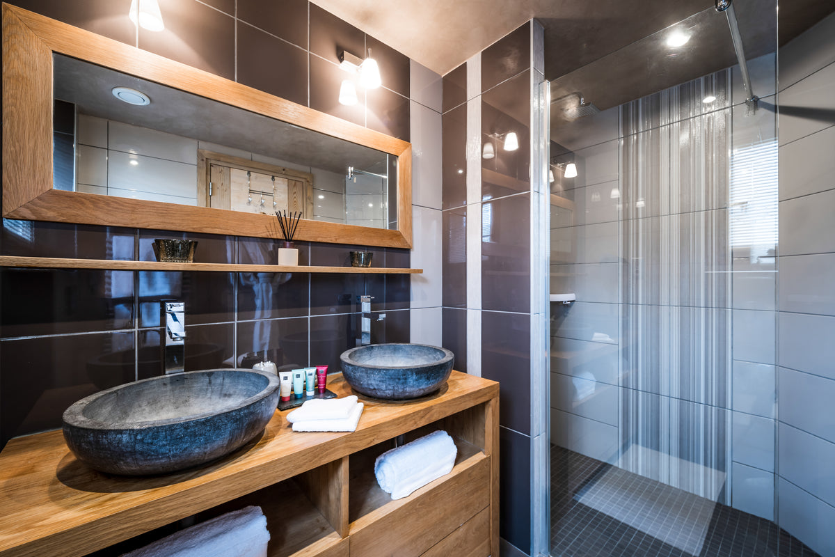 Modern bathroom interior with a double vessel sink setup on a wooden vanity, a large wood-framed mirror, dark wall tiles, and a glass shower enclosure with a sheer curtain.