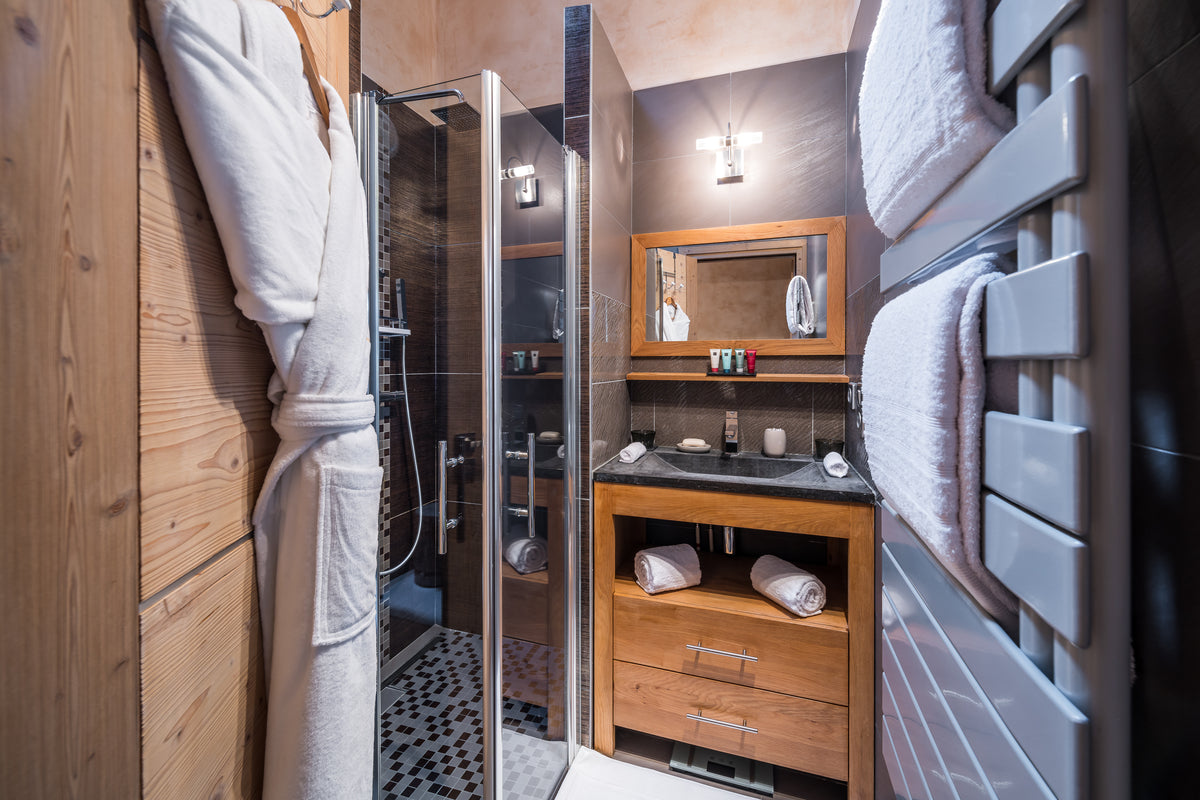 A modern luxury bathroom interior featuring a walk-in glass shower enclosure, a wooden vanity with a dark countertop, a mirror, and a towel rail holding white towels.