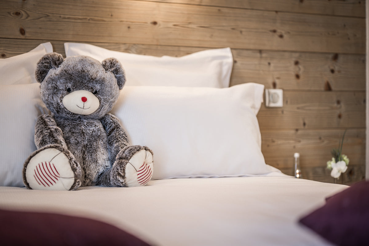 A plush gray teddy bear with white and red striped feet sits on a neatly made bed with white pillows and sheets, set against a rustic wooden headboard.