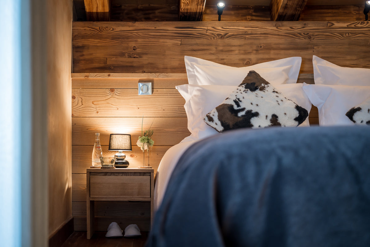 A cozy bedroom scene with a bed dressed in white linens and a black-and-white cowhide-pattern pillow, set against a rustic wood-paneled wall. A wooden bedside table with a lit lamp, bottle, and small plant sits beside the bed, and a dark blue blanket is visible in the foreground.