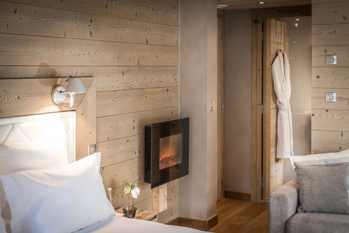 A cozy modern bedroom interior with light wood paneled walls, a built-in fireplace, white bedding, a gray armchair, and a robe hanging in the background.