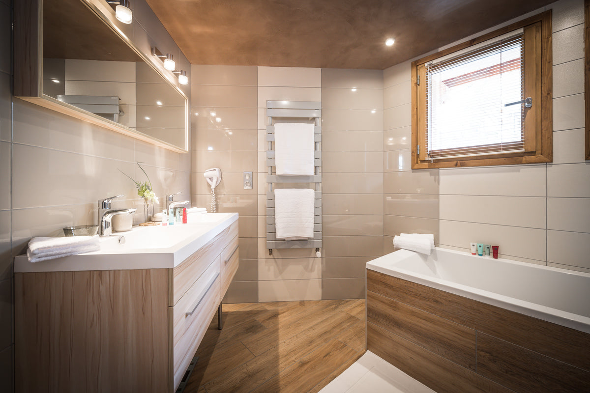 Modern bathroom interior with a double vanity, large mirror, heated towel rail, bathtub with wood paneling, wood floor, and beige tiled walls.