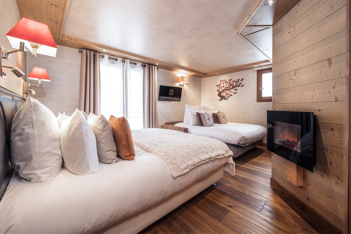 A cozy mountain-style bedroom with two neatly made beds, white bedding, red wall lamps, wood-paneled walls, and a built-in fireplace.