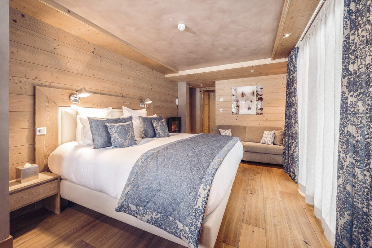 A luxury bedroom interior with a large bed dressed in white linens and a blue patterned throw blanket, set against wood-paneled walls and a wood floor. Matching blue patterned curtains hang along the right side, and a sofa is visible in the background.