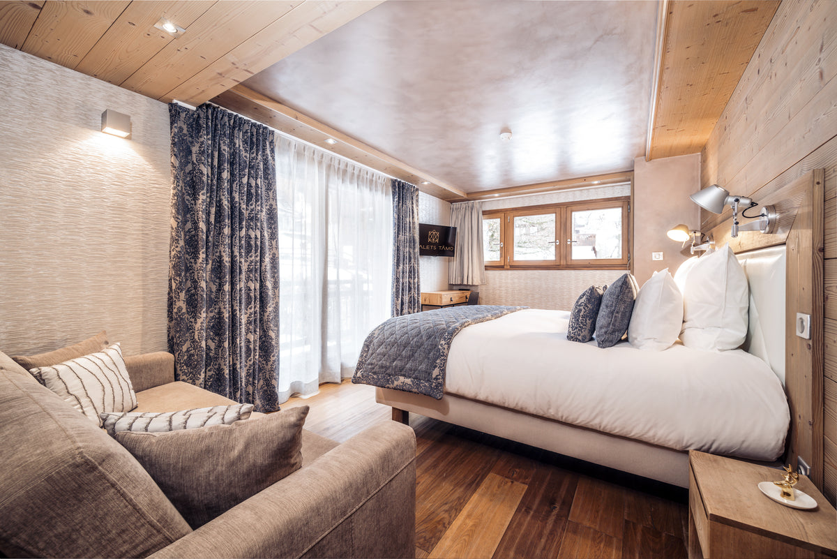 A modern ski chalet bedroom featuring a large bed with white linens and blue-gray pillows, a matching armchair in the foreground, patterned blue curtains, and light wood walls and ceiling.