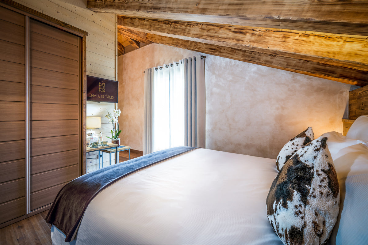 A rustic bedroom interior with a large bed dressed in white linens and a dark throw blanket, set under a sloped wooden ceiling with exposed beams. A patterned pillow rests on the bed, and a window with sheer curtains is visible in the background.