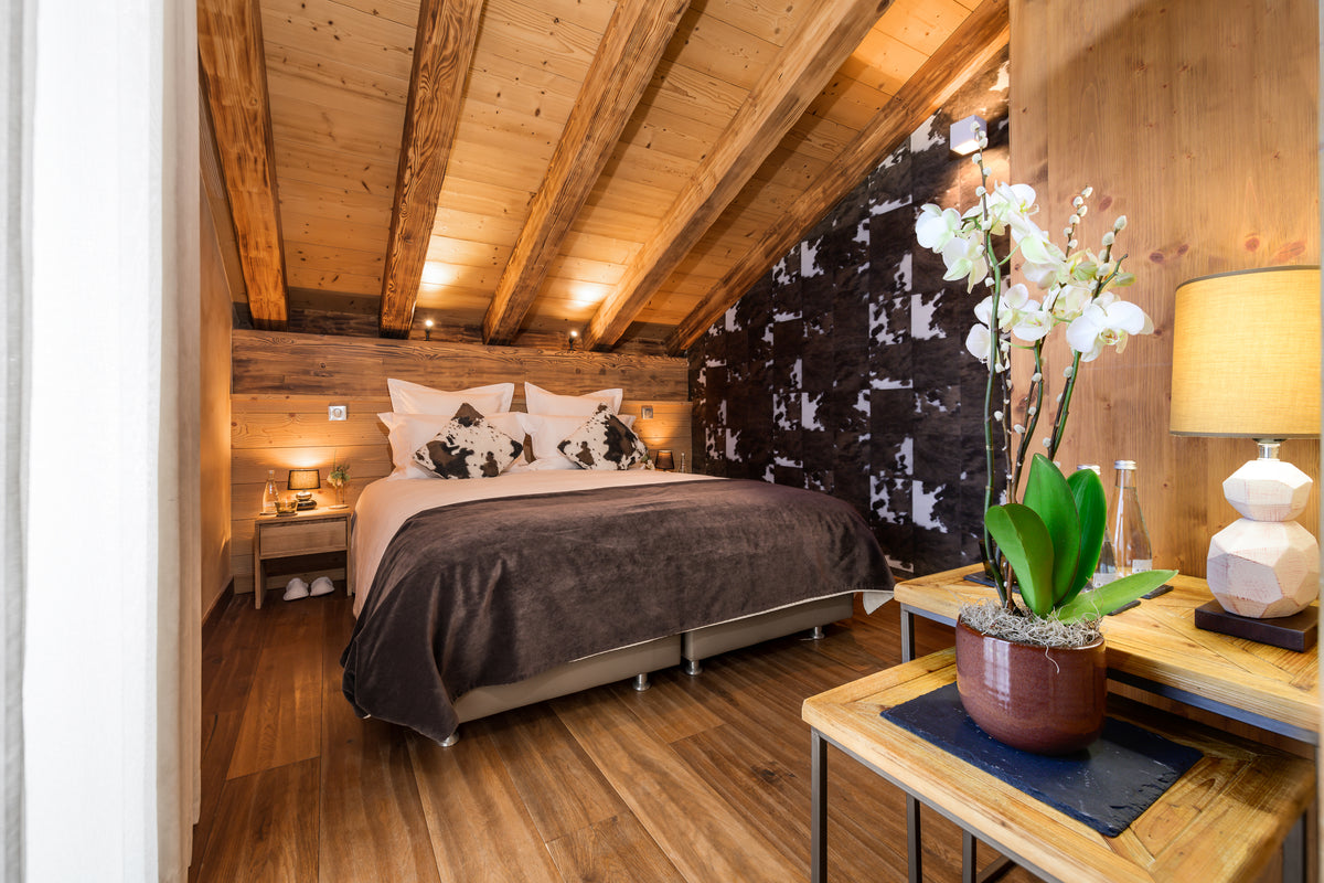 A cozy bedroom interior with a bed dressed in white and dark brown bedding, set against a wood-paneled wall and slanted wooden ceiling. A black-and-white patterned accent wall is visible behind the bed, and a wooden table in the foreground holds a potted white orchid and a lit table lamp.