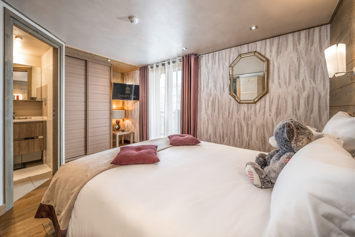 A modern bedroom interior featuring a large bed with white linens, two dusty rose pillows, a plush stuffed animal on the pillow, patterned wallpaper, a gold-framed octagonal mirror, and an open wardrobe door revealing a bathroom.