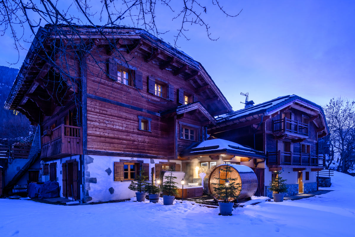 A large traditional alpine chalet with wooden walls and snow-covered roofs, lit warmly at dusk in a snowy mountain setting.