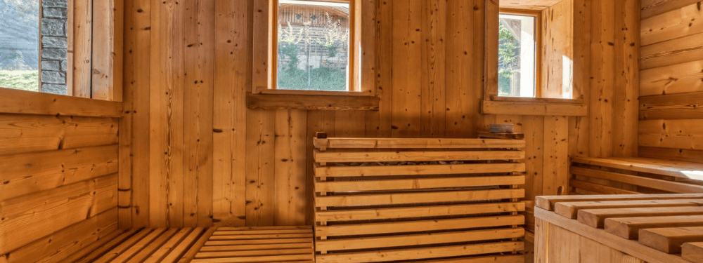 A wooden sauna interior with light brown wood-paneled walls, two windows, a wooden bench in the foreground, and a wooden staircase or seating area on the right.