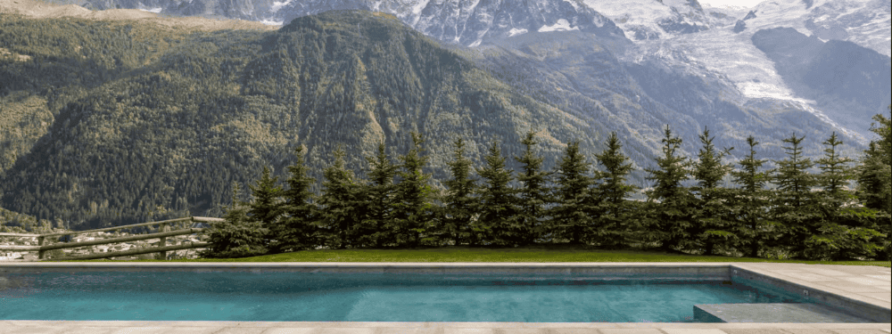 A luxury outdoor swimming pool with clear blue water in the foreground, bordered by a neat green lawn and a row of dark pine trees, with majestic snow-capped mountains and forested slopes in the background.