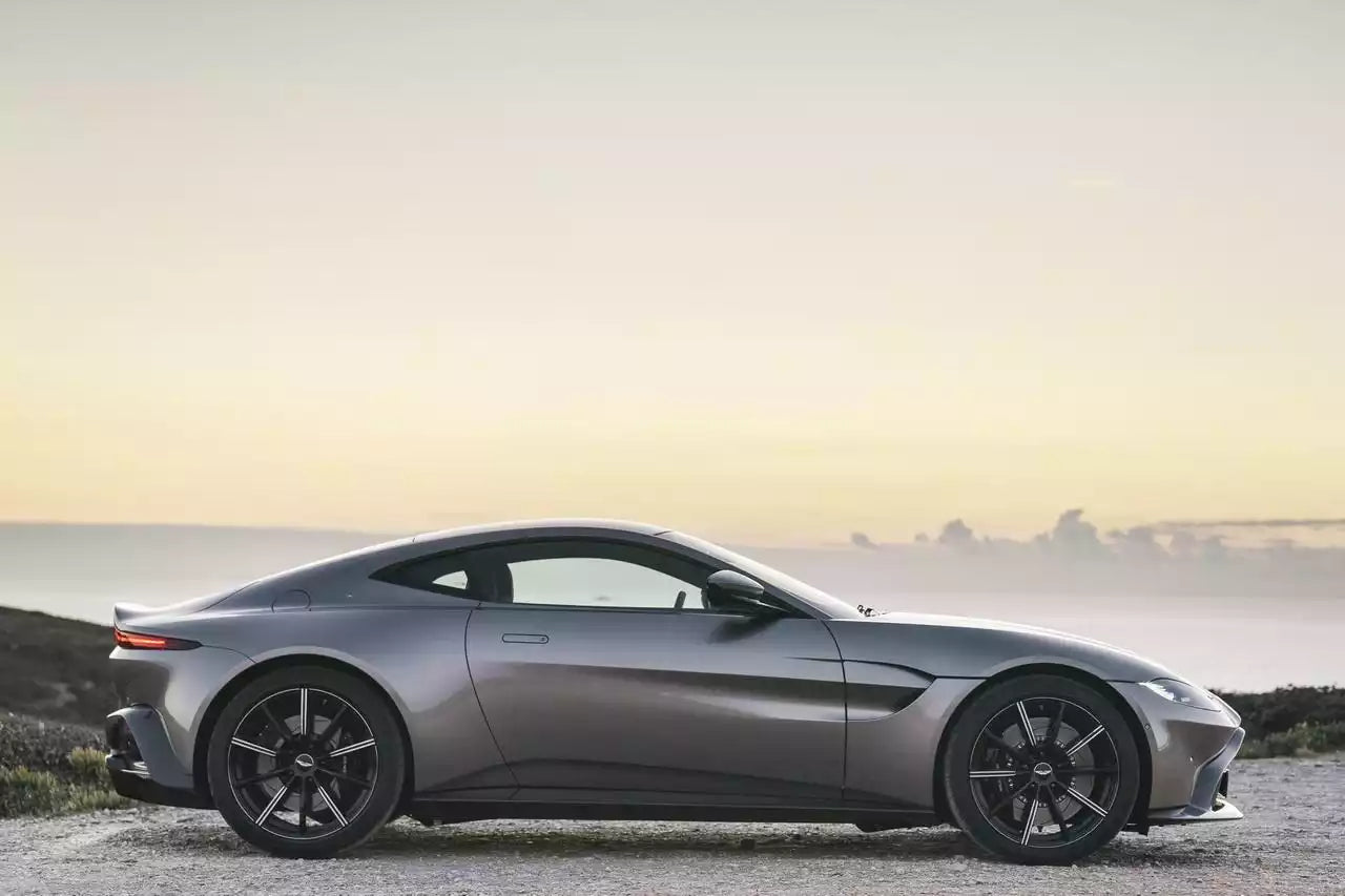 A dark gray Aston Martin sports car shown in a side profile view, parked outdoors on a gravel surface with a hazy coastal landscape in the background.