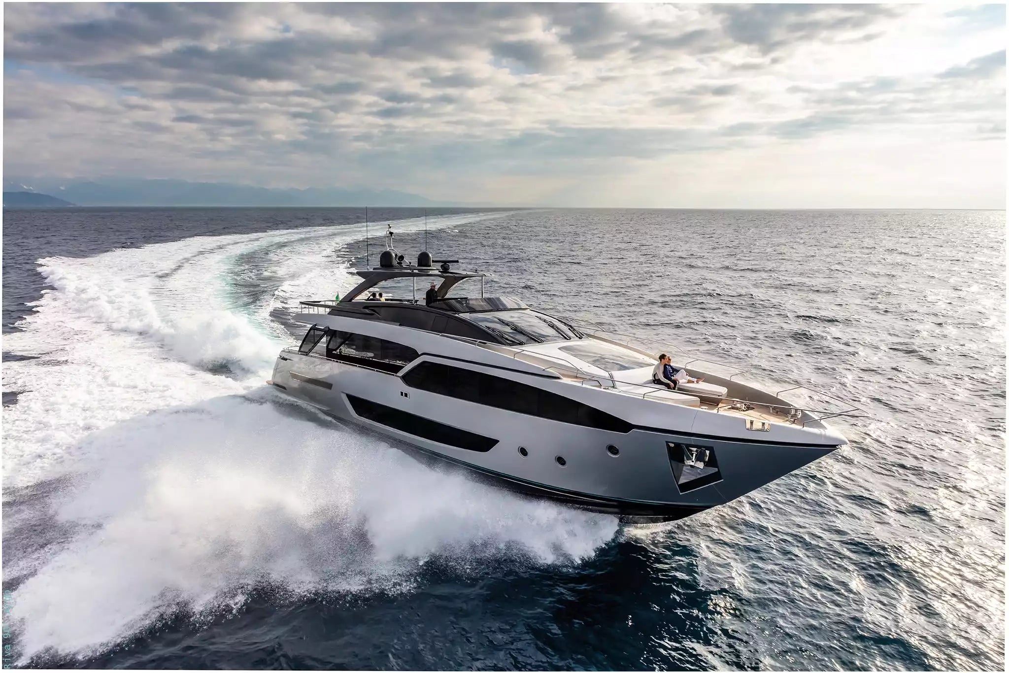 A luxury motor yacht speeds across open ocean water, creating a large white wake. The yacht is white with dark gray and black accents, photographed from an elevated angle under a cloudy sky.