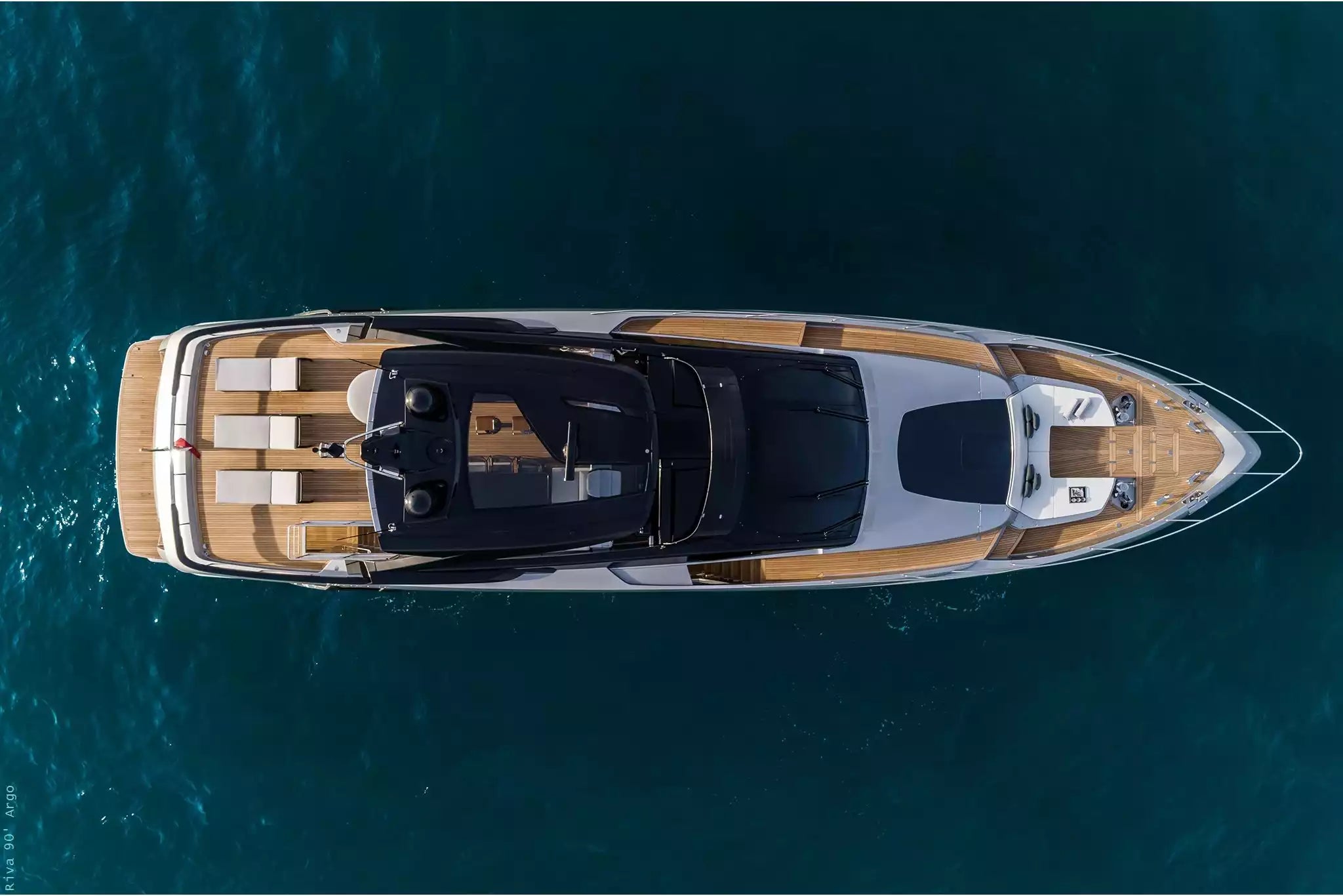 Overhead view of a luxury yacht floating on deep blue ocean water, featuring a white hull, warm wood-toned deck surfaces, and a black upper deck canopy.