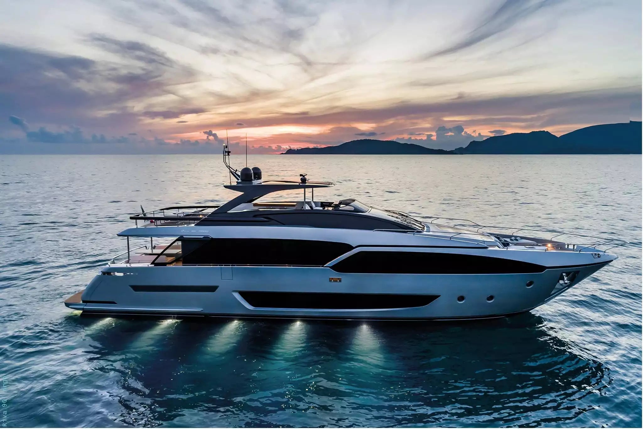 A luxury motor yacht with a white hull and dark gray superstructure, sailing on calm blue water during a dramatic sunset with clouds and distant islands.