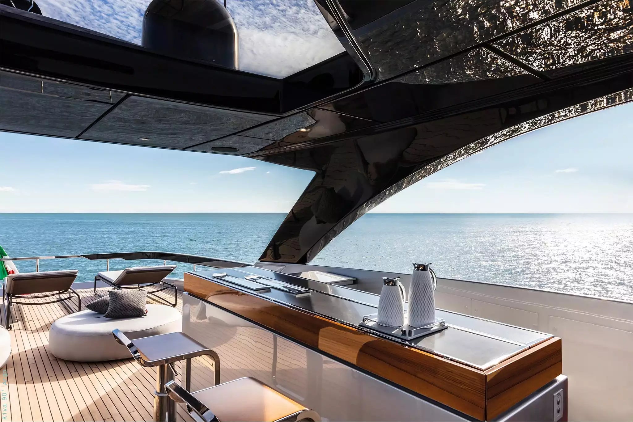 A luxury yacht outdoor bar and lounge area with a wooden counter, stainless steel sink, two white pitchers, lounge chairs, and an open view of the blue ocean under a bright sky.