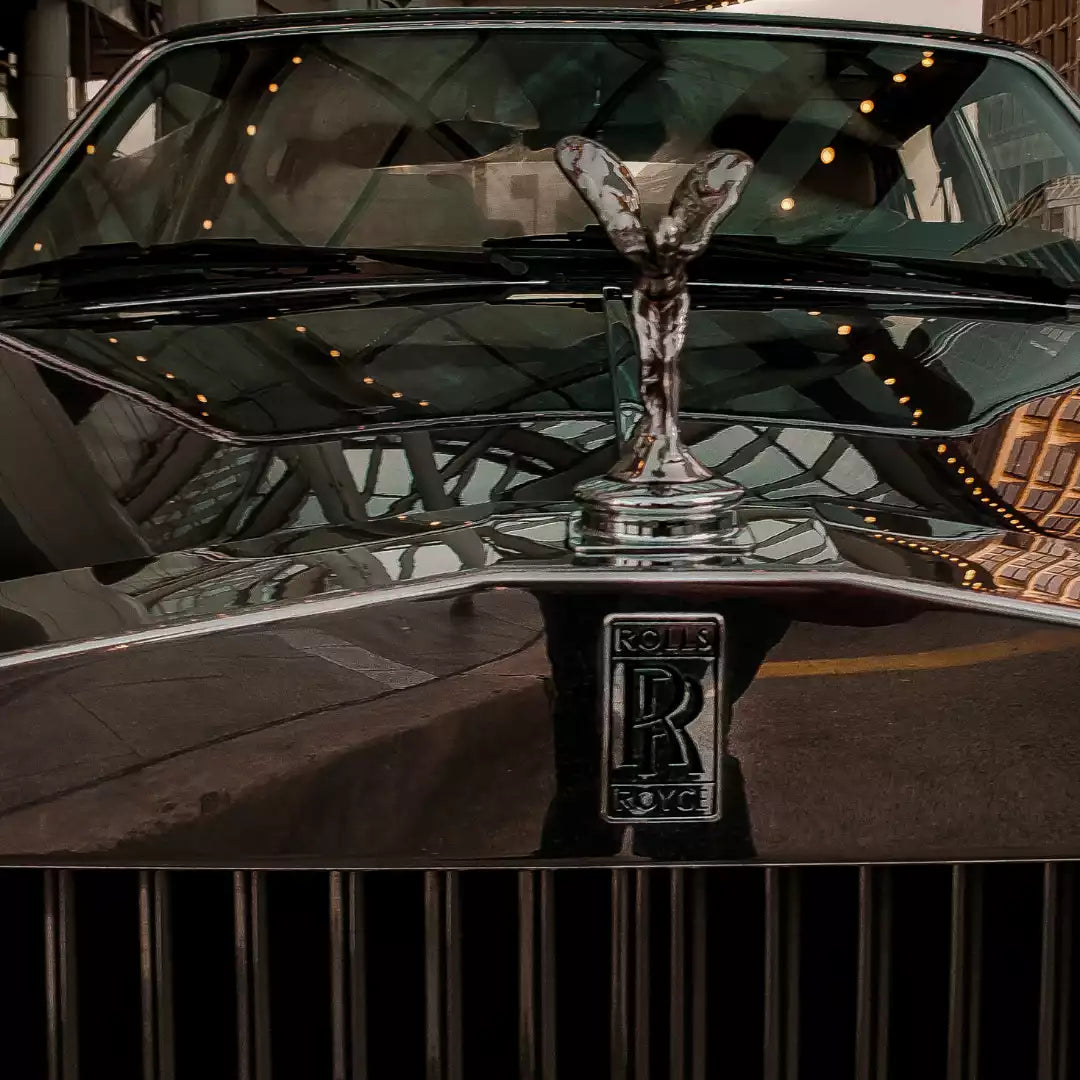 Close-up of the front hood and grille of a Rolls-Royce car, featuring the chrome Spirit of Rolls-Royce hood ornament and the Rolls-Royce badge on a glossy black finish.