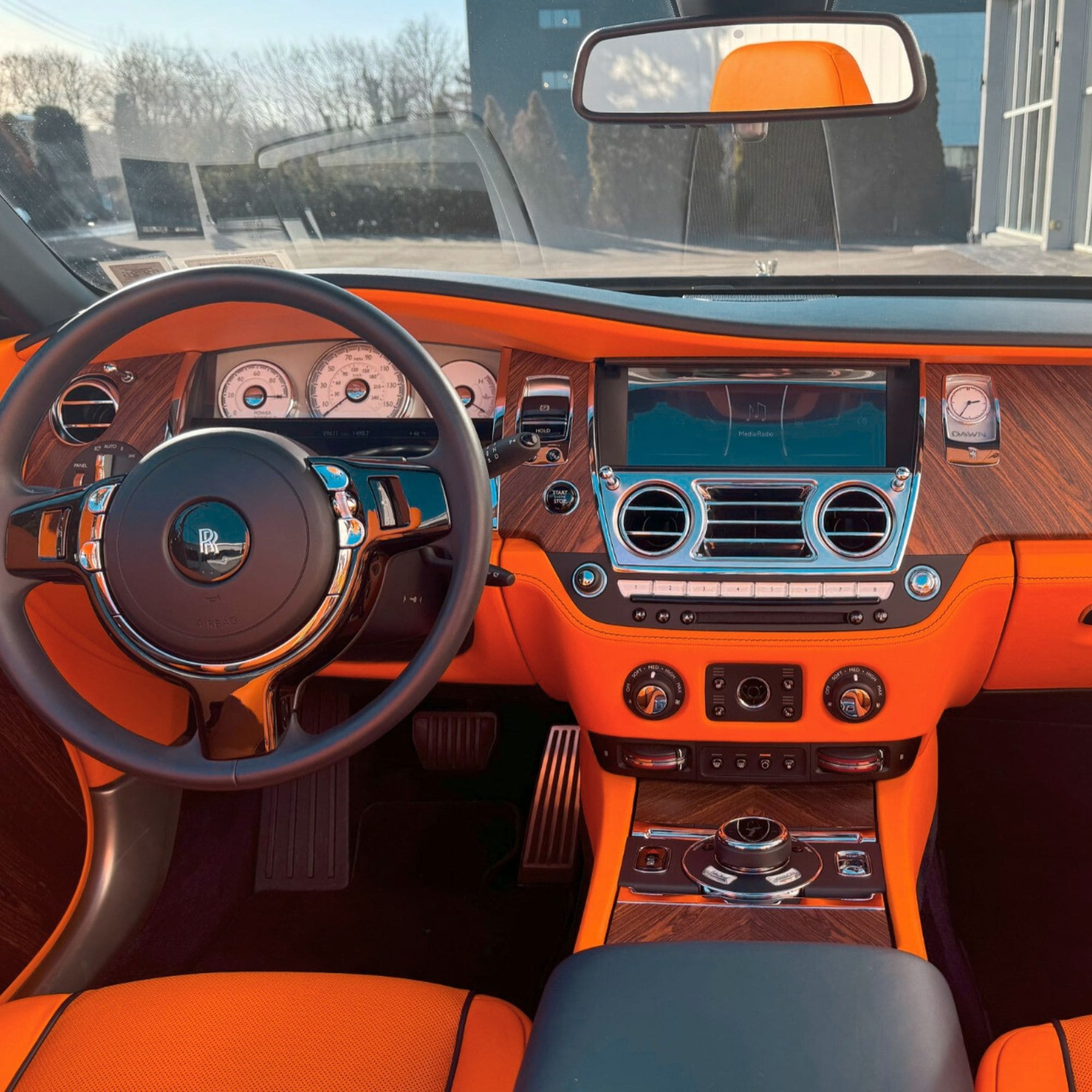 Luxury car interior with bright orange leather seats and matching orange door panels, dark wood trim, a black steering wheel with a Rolls-Royce logo, and a center console with a touchscreen display.