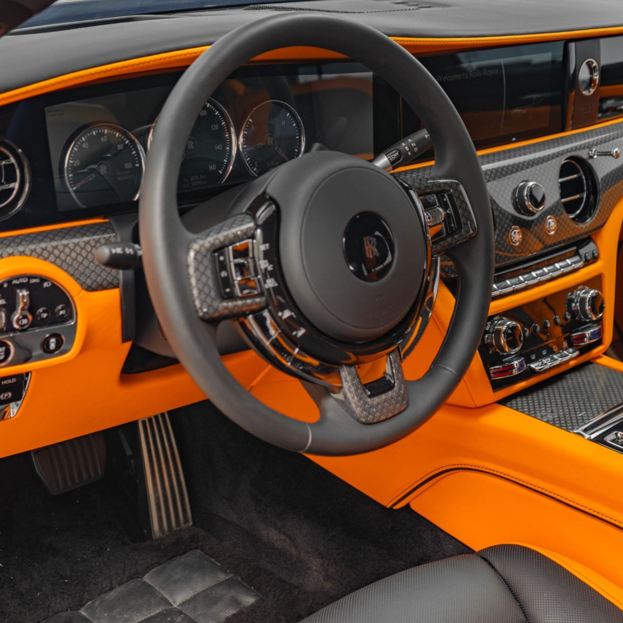Close-up of a luxury car interior featuring a black leather steering wheel with carbon fiber accents, black dashboard, and vivid orange trim panels.