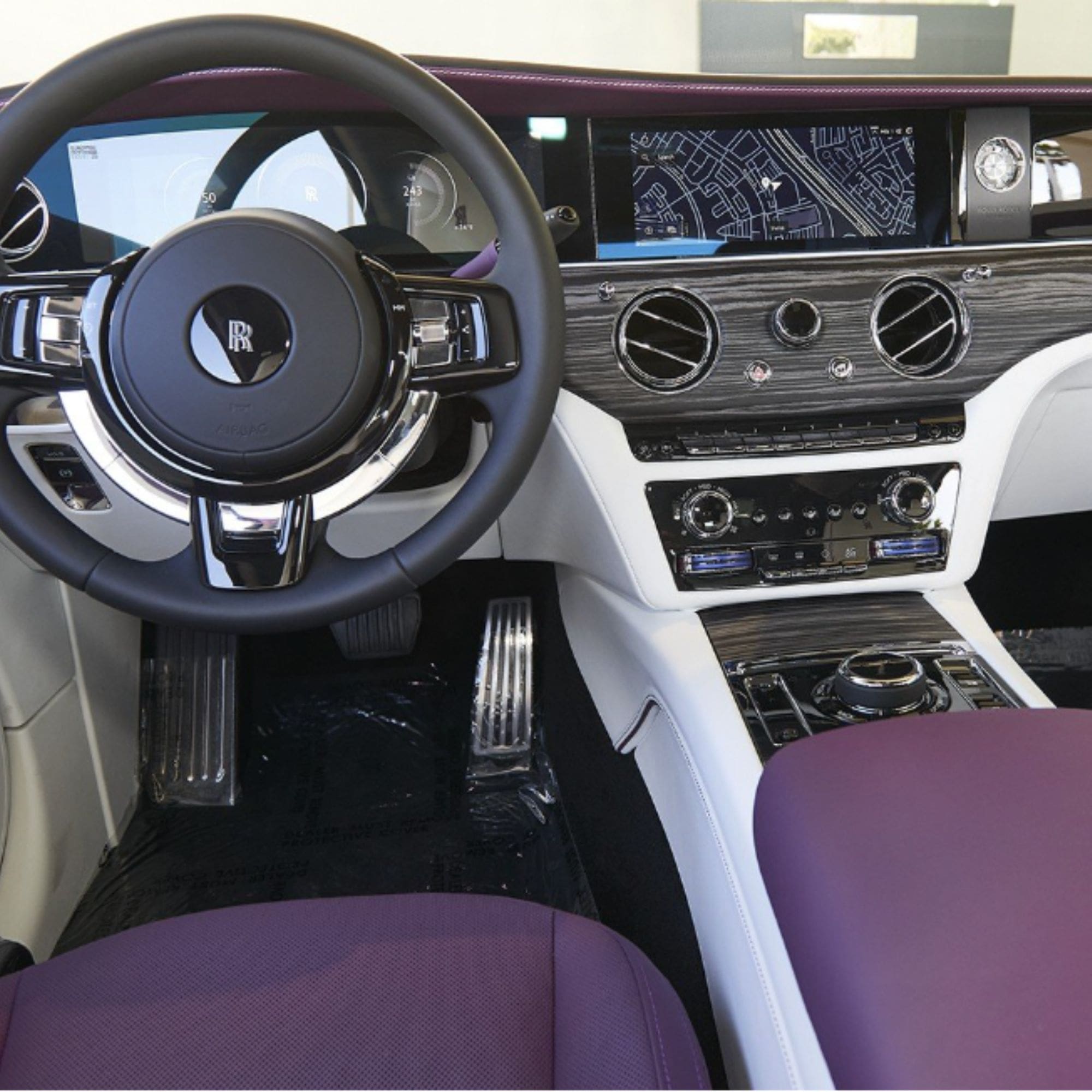 Luxury car interior featuring a black leather steering wheel with a silver and black Rolls-Royce logo, a digital dashboard display, wood trim, and purple leather seats.