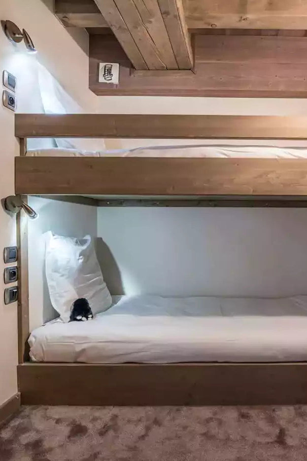 A compact wooden bunk bed with white bedding in a cozy, compact room with warm lighting and textured walls.