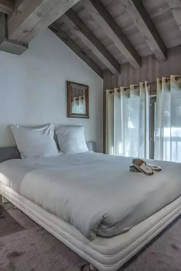 A modern bedroom with a large bed dressed in white bedding, set in a rustic room with exposed wooden ceiling beams, light walls, sheer curtains, and a wooden cabinet in the foreground.