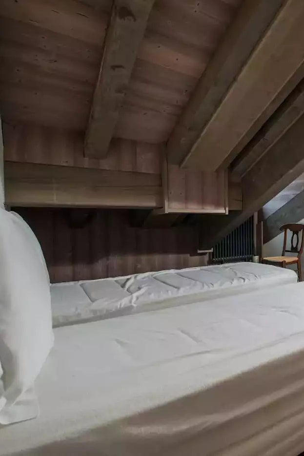 A cozy attic bedroom with two single beds made from a larger bed frame, dressed in white linens. The room features a rustic wooden slatted ceiling, a purple carpet, and a dark wood chair with a woven seat cushion against a light-colored wall.