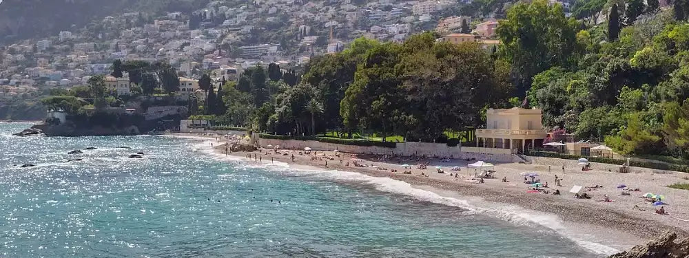 A scenic coastal landscape with turquoise water, a sandy beach with small beachgoers, dense green trees, and a hillside town in the background.
