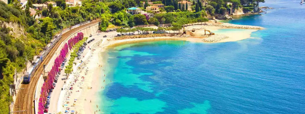 Aerial view of a sunny coastal beach with turquoise water, pale sand, a curved shoreline lined with pink flower-covered railings, and green hillside buildings above.