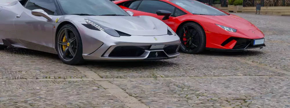 Two luxury sports cars, one silver and one red, on a cobblestone street.
