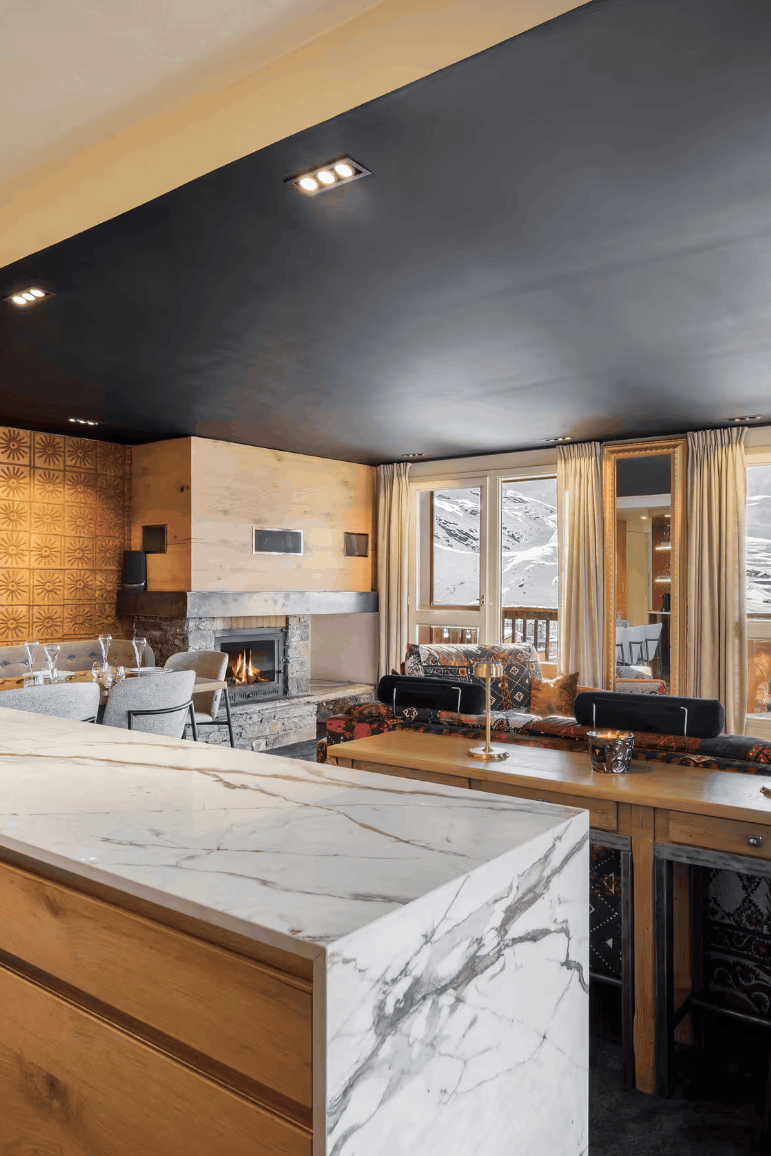 A luxury modern ski chalet interior featuring a large marble-topped kitchen island with wood grain cabinets, a dark ceiling, a dining area, a home office desk setup, and large windows overlooking a mountain view.