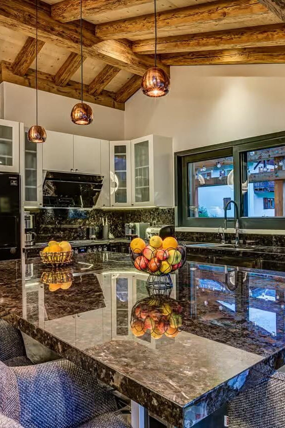 A luxury modern kitchen with a large dark marble island, textured gray upholstered bar stools, rustic wooden ceiling beams, and black stainless steel appliances.