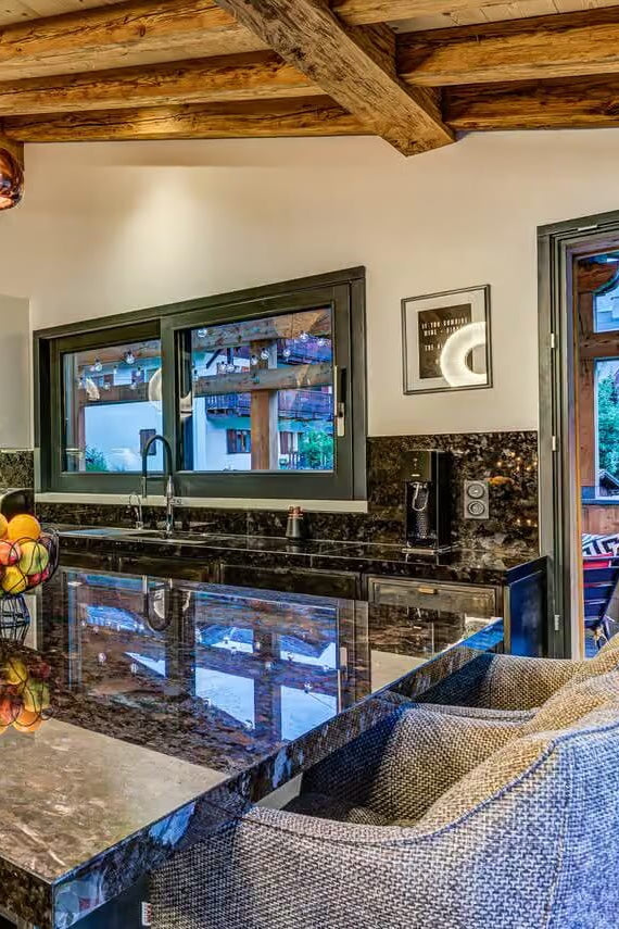 A luxury modern kitchen with a large dark marble island, textured gray upholstered bar stools, rustic wooden ceiling beams, and black stainless steel appliances.