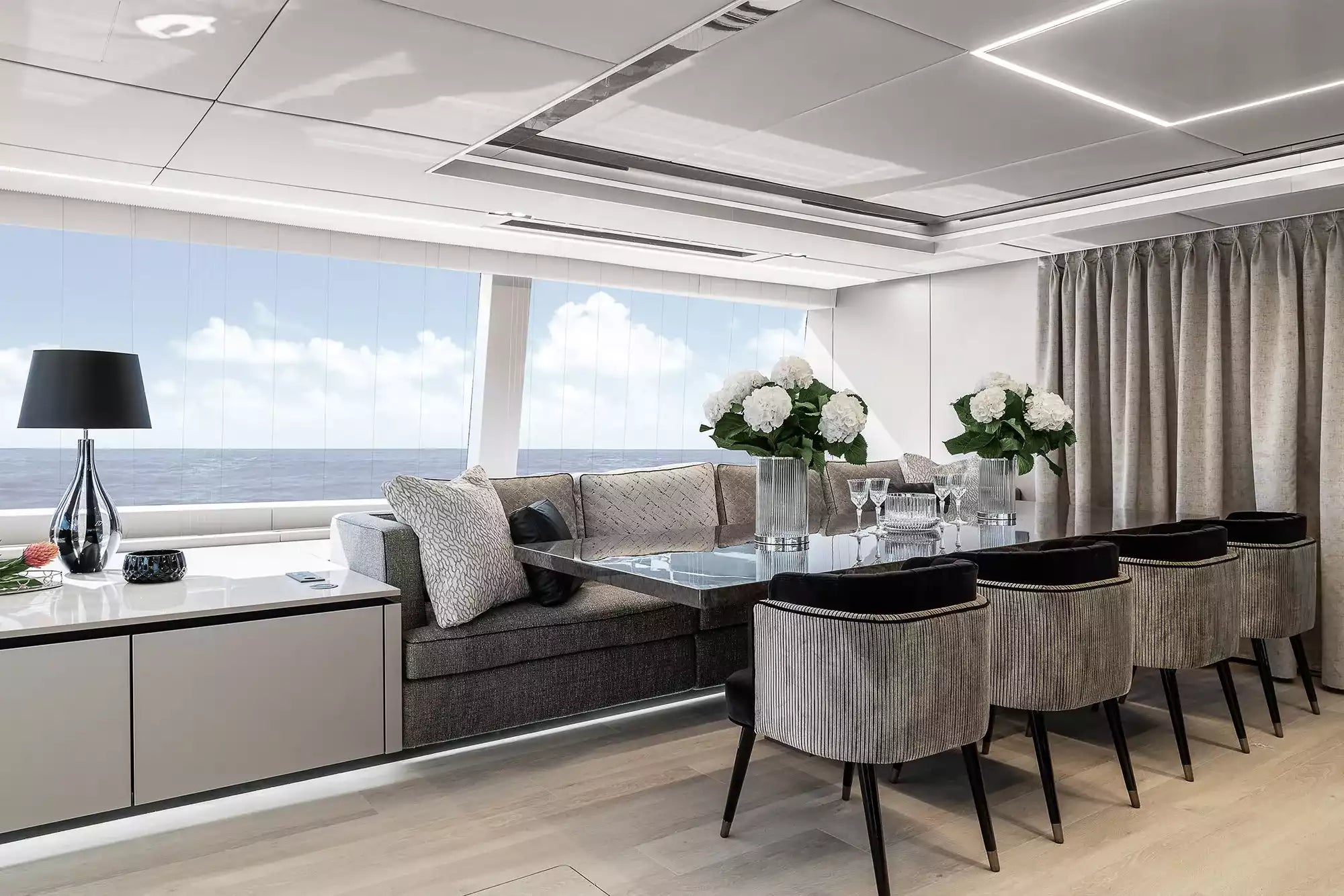 A luxury yacht or modern villa interior featuring a dining area with a glass-top table, six upholstered gray chairs, a gray sofa with patterned cushions, and a large window overlooking the ocean.