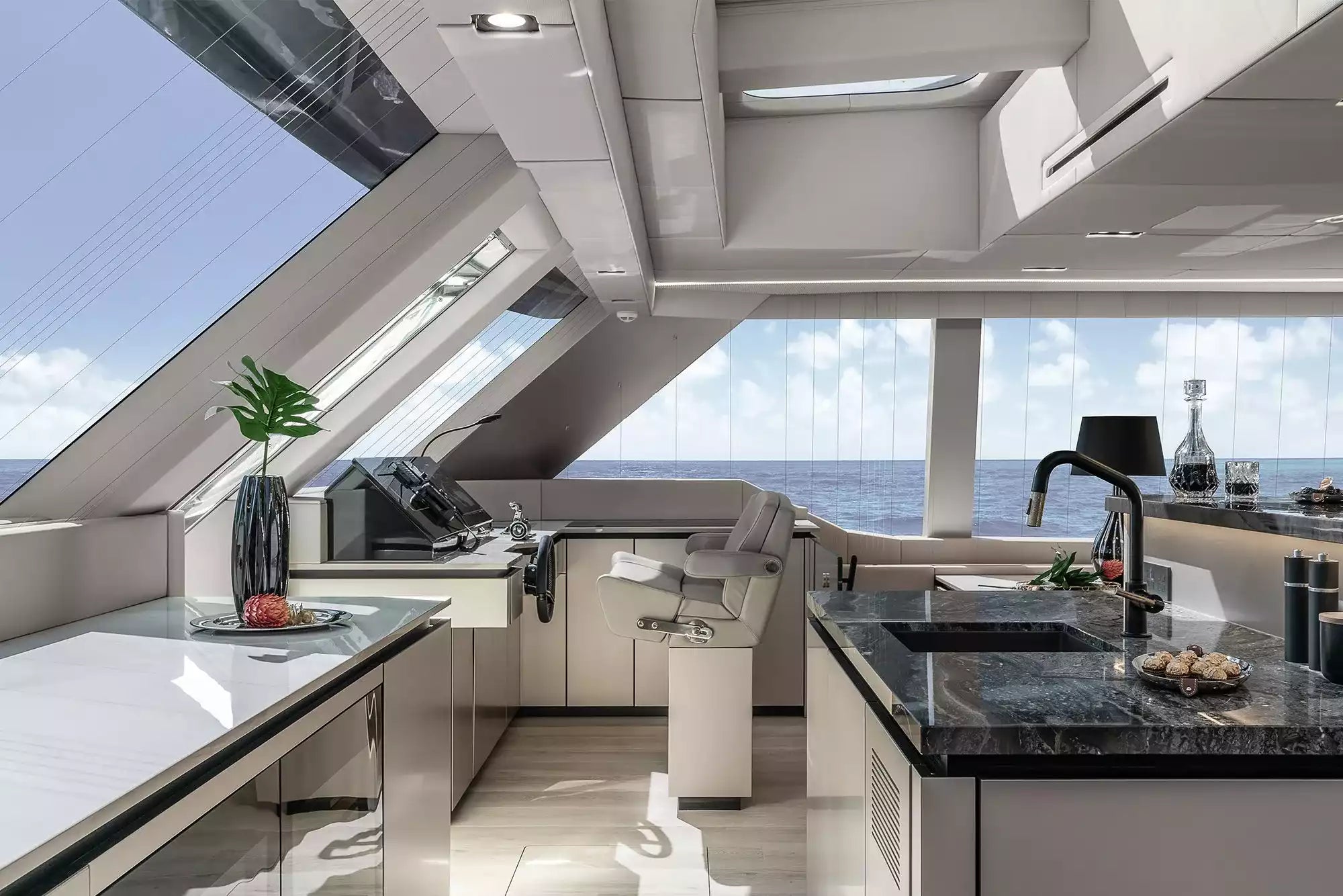 A luxury yacht galley kitchen featuring modern white cabinetry, dark marble countertops, a central sink, and a large view of the open ocean through the windows.