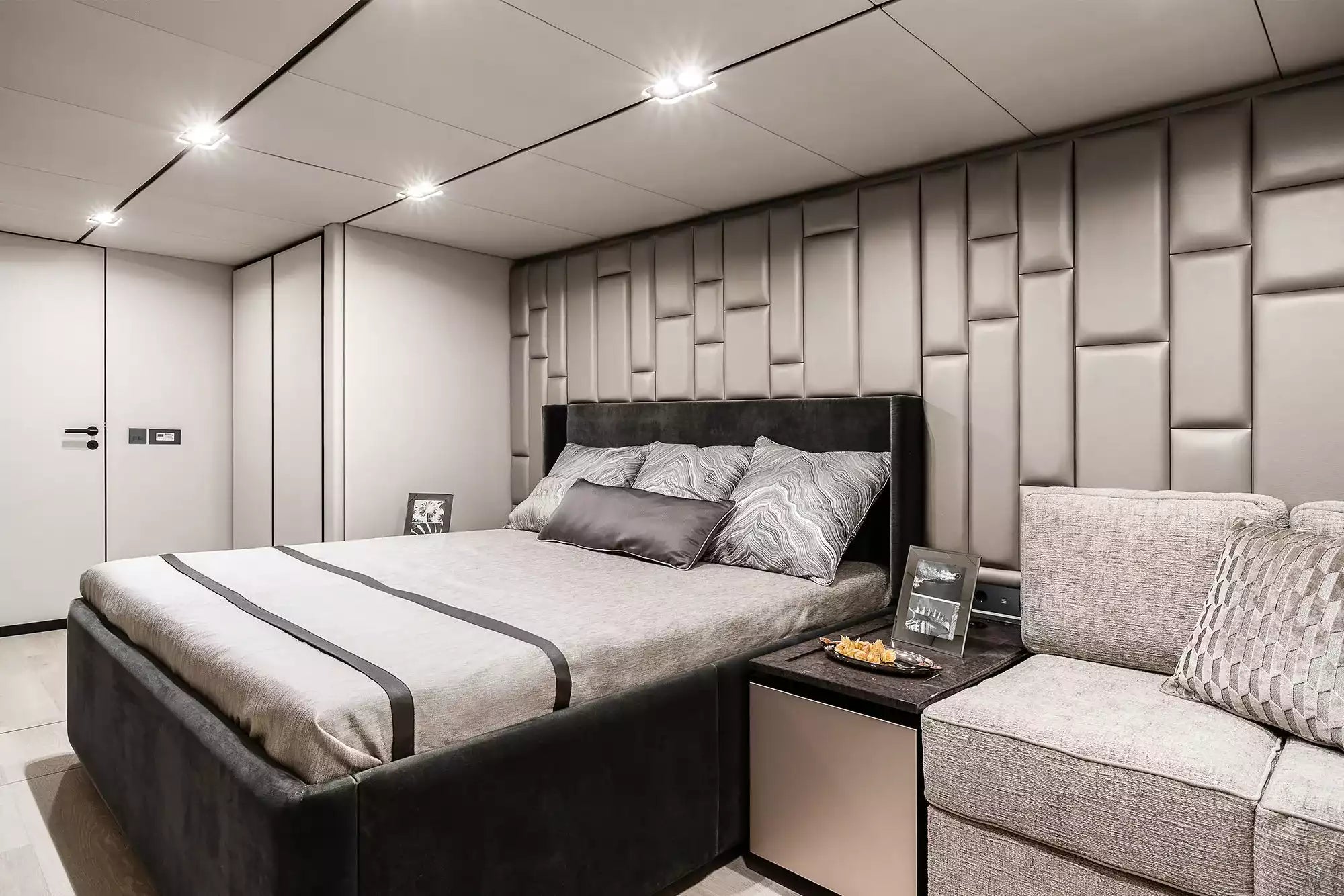 A modern bedroom interior featuring a bed with gray and black bedding, a textured gray upholstered headboard and side table, a matching sofa, and a paneled gray accent wall under recessed ceiling lights.