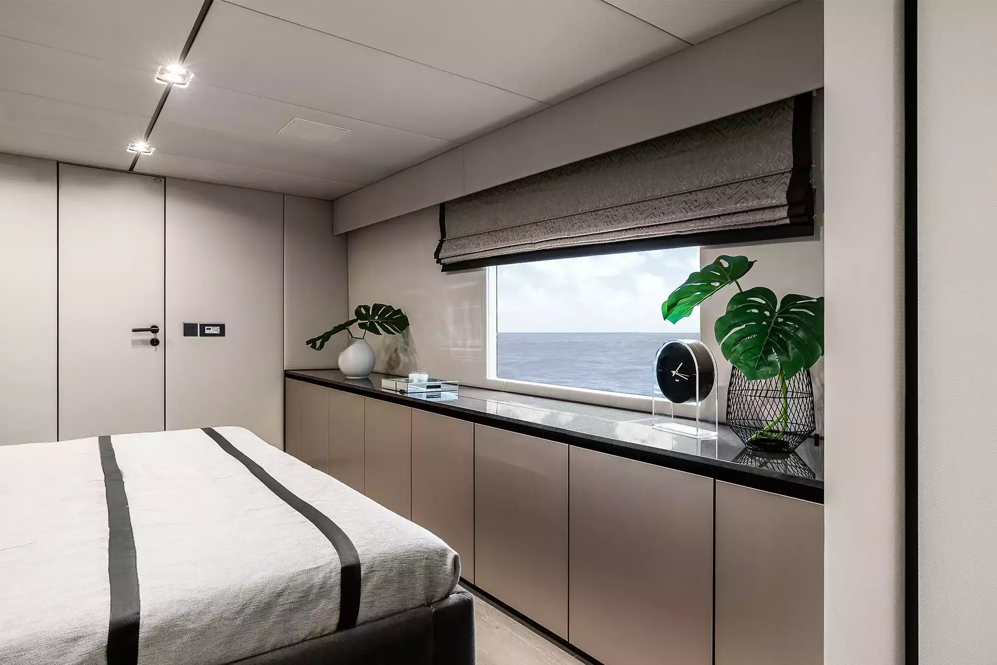A modern yacht or luxury bedroom interior featuring a bed with white and black striped bedding, sleek metallic cabinetry, a black countertop, and a large window overlooking the ocean.