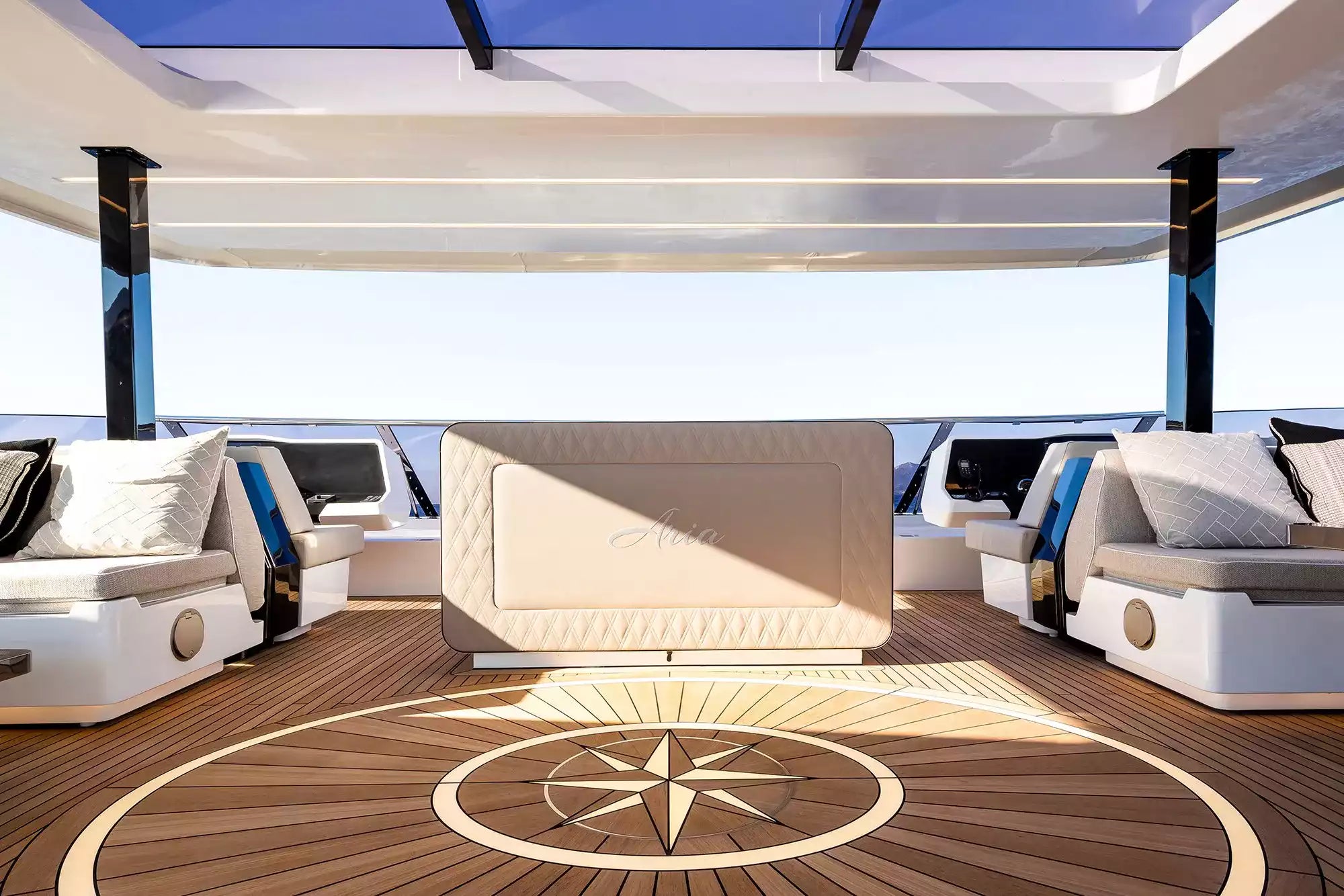 Luxury yacht deck lounge featuring a central cream tufted sofa, white daybeds with blue accents on both sides, a wooden teak deck, and a circular compass rose inlay under a glass canopy.