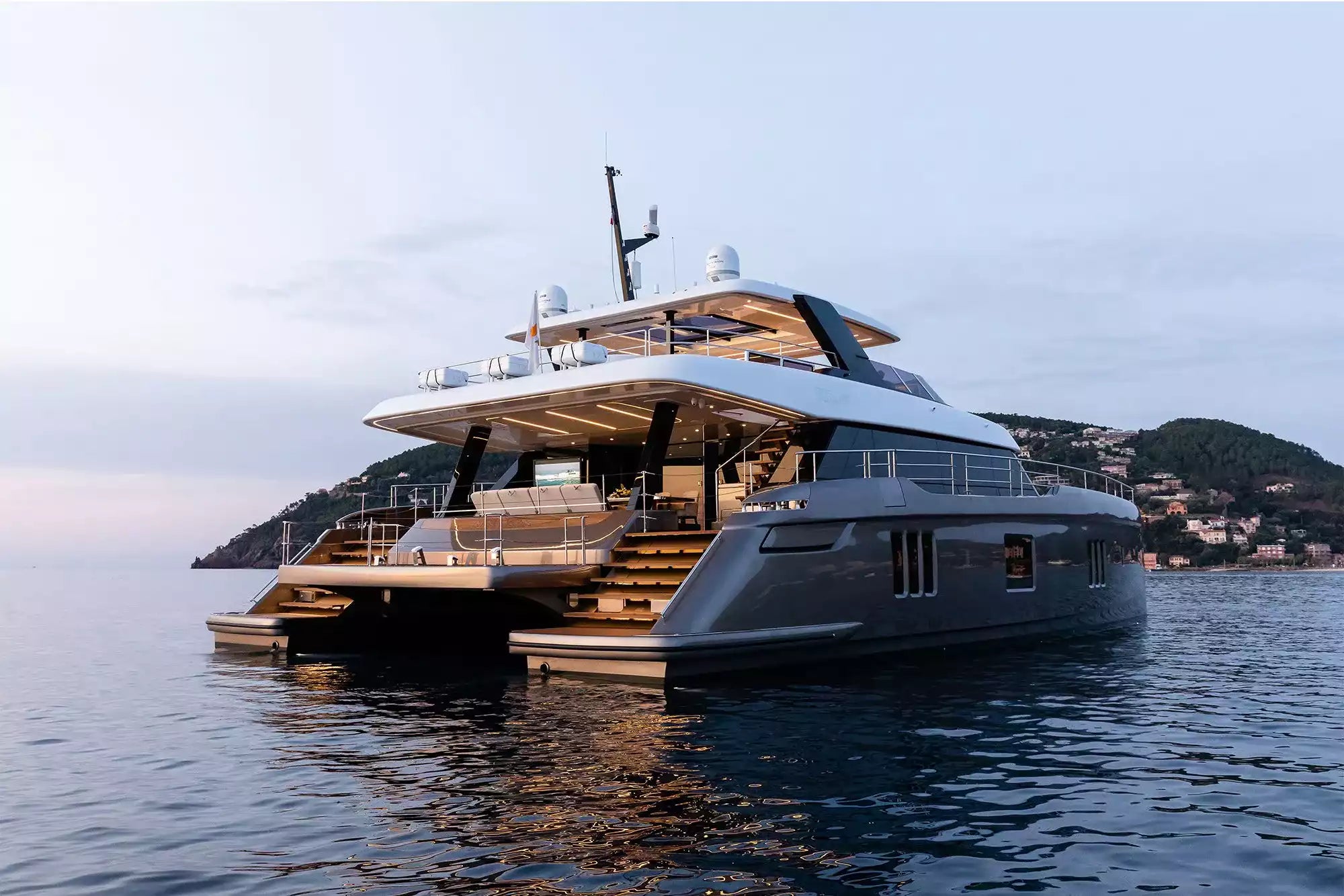 A luxury yacht with a dark gray hull and white superstructure floating on calm water, with a hillside village visible in the background under a soft sky.