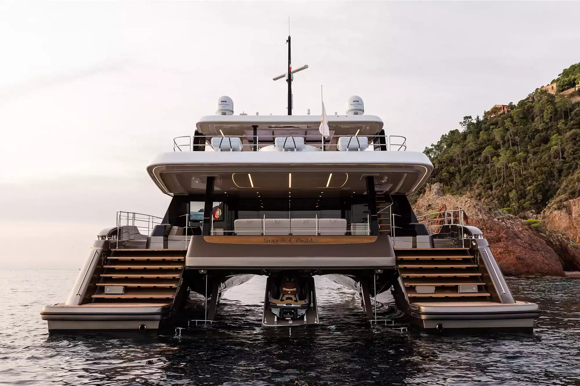 A luxury superyacht shown from the rear, floating on water with a rocky, forested cliff on the right and an overcast sky above. The yacht features a white superstructure, dark wood accents, and dual stairways leading to outdoor decks.