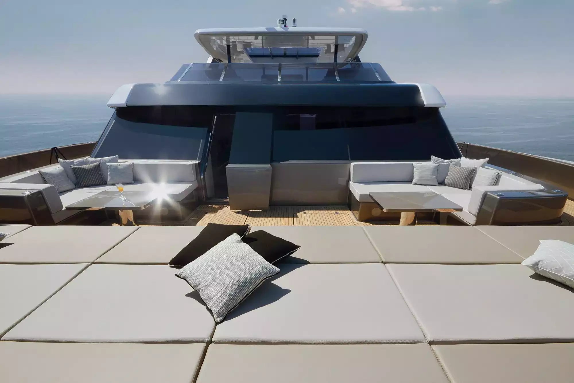 A luxury yacht deck with outdoor lounge seating, cushions, and a sun deck overlooking the ocean under a clear sky.